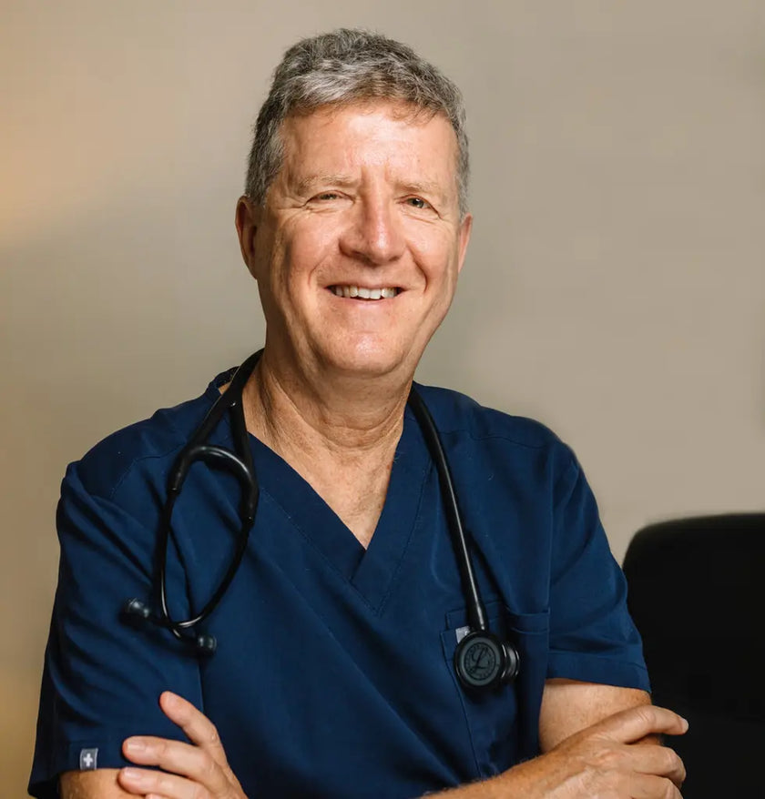 Dr Isaac Eliaz wearing blue scrubs with a stethoscope around his neck, smiling against a neutral background