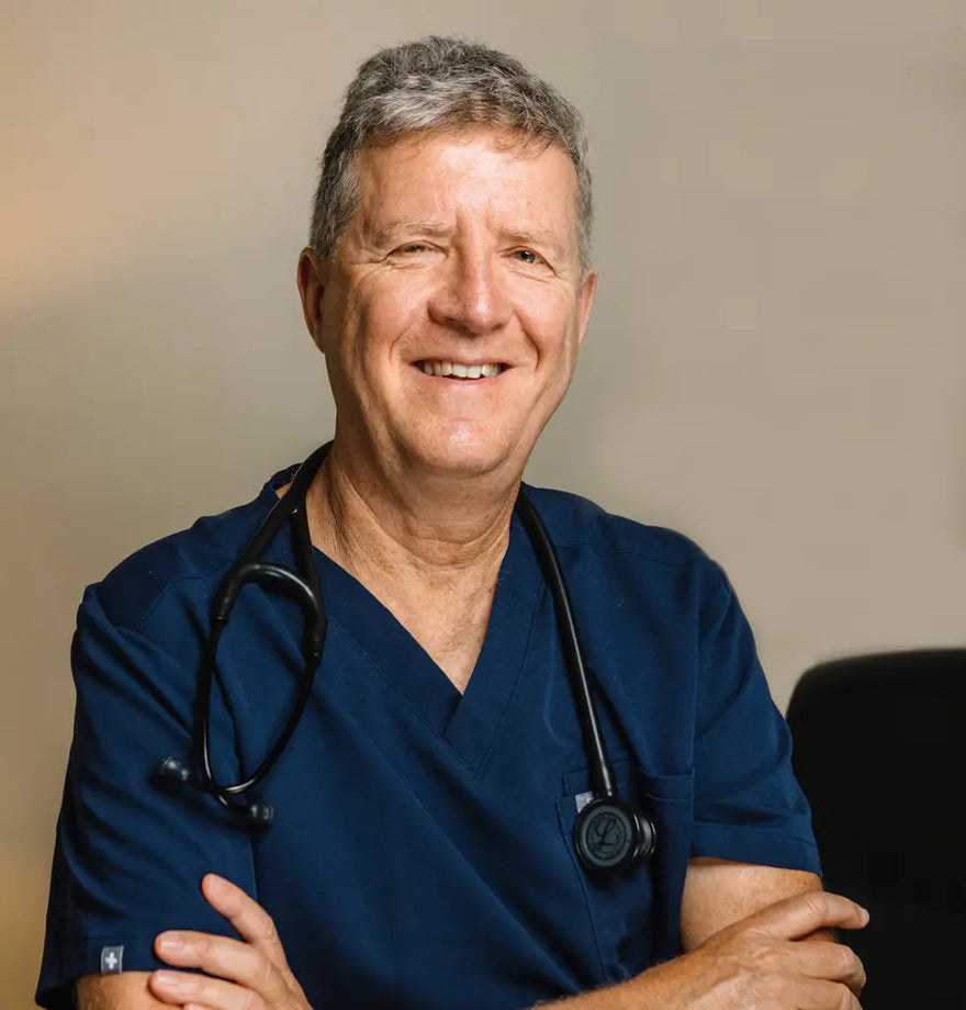 Dr Isaac Eliaz wearing blue scrubs with a stethoscope around his neck, smiling against a neutral background