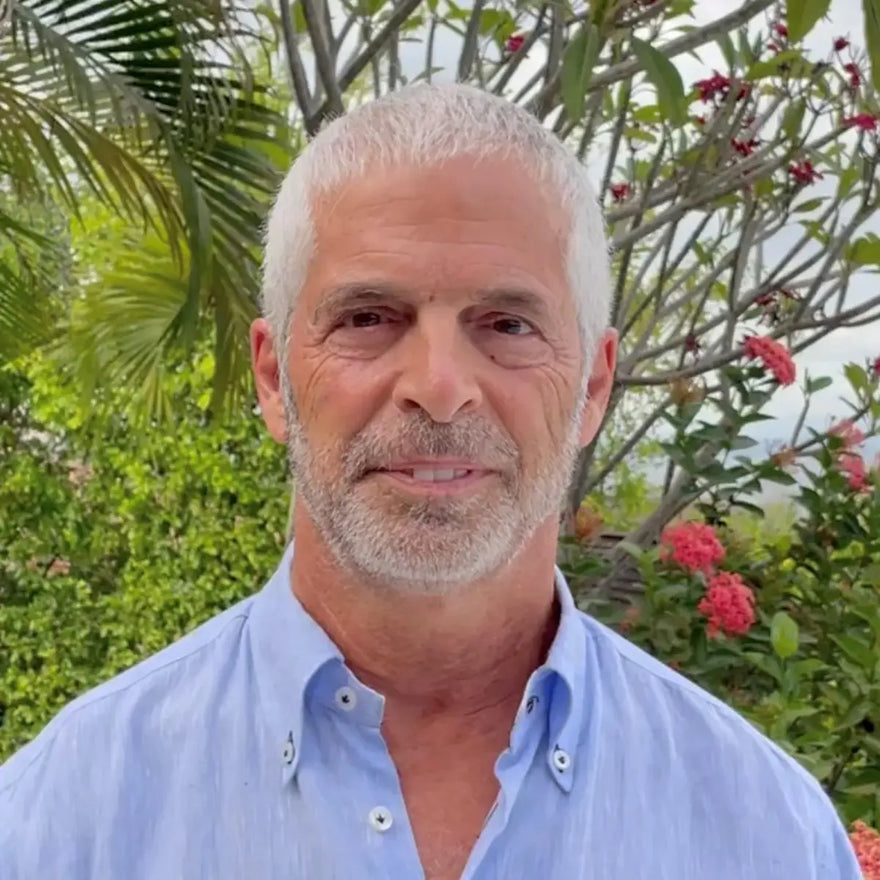 Dr Tom O'Bryan wearing a light blue shirt outdoors with greenery and flowers in the background